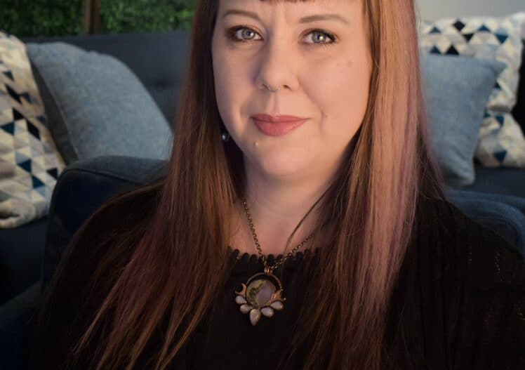 Headshot of Asta MacBaine, Manager of Operations Services & Training, sitting on a blue couch dressed in a dark top, wearing a large pendant necklace.
