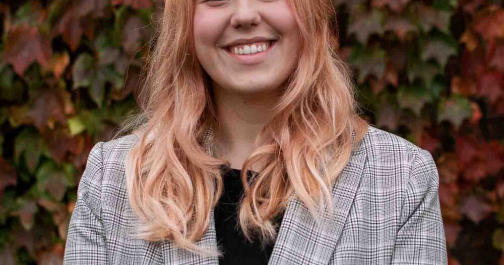 Headshot of a smiling woman with blond hair wearing a dark top and striped business jacket
