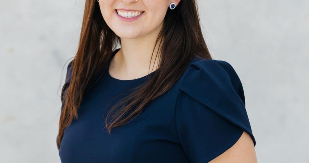 Headshot of a woman with long brown hair dressed in a dark blue top wearing blue pendant earrings