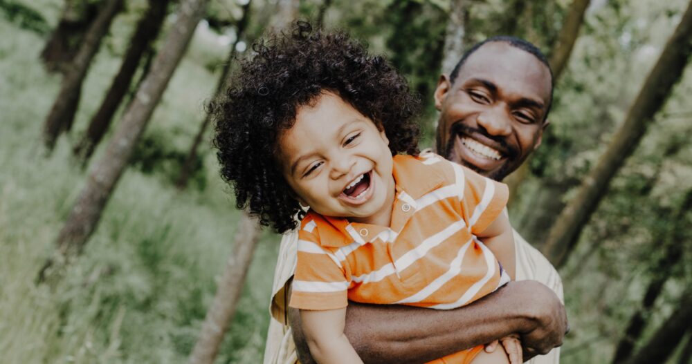 A young boy dressed in an orange and white polo shirt being held by his smiling father against a backdrop of trees and greenery.