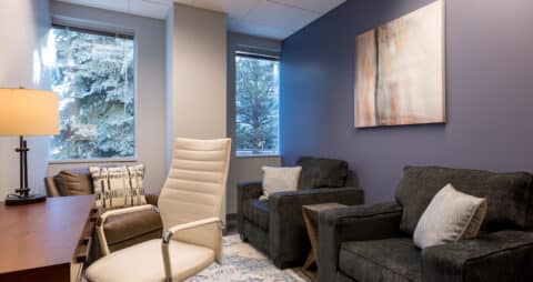 An interior photo of an Ellie Mental Health Therapy Clinic office with two dark gray chairs, a brown leather chair, a small desk with desk lamp and white office chair, and an abstract painting hanging on one wall.