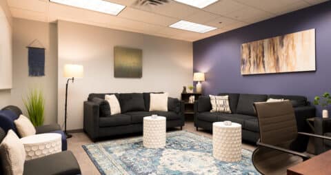 An interior photo of an Ellie Mental Health Therapy Clinic waiting area that has one of the walls painted purple with dark colored couches and chairs, small white lamps, and paintings on the walls.
