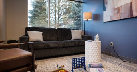 An interior photo of an Ellie Mental Health Therapy Clinic office with a large window, a dark gray sofa, a tan leather chair, a small white table and an abstract painting hanging on a blue wall.