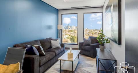 An interior photo of an Ellie Mental Health Therapy Clinic office with two large windows, a gray sofa and chair, a table lamp, small white table and a wooden bookcase.