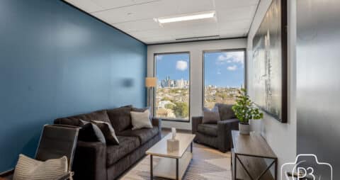 An interior photo of an Ellie Mental Health Therapy Clinic office with two large windows, a gray sofa and chair, a table lamp, small white table and a wooden bookcase.