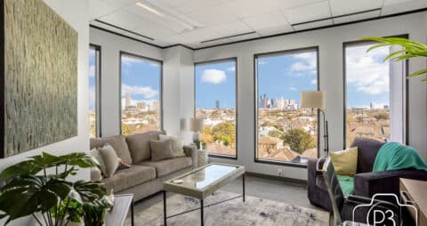 An interior photo of an Ellie Mental Health Therapy Clinic office with 5 large windows, a large gray couch, a single dark gray chair, with various plants and a large painting hanging on one wall.