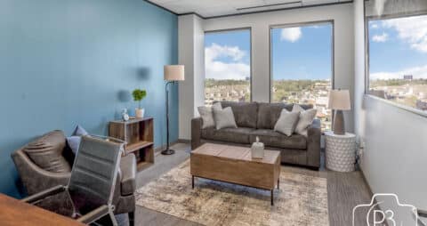 An interior photo of an Ellie Mental Health Therapy Clinic office with two large windows, a gray couch, a table lamp and floor lamp, a brown leather chair, and a small desk and white office chair.