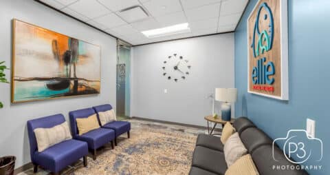 An interior photo of an Ellie Mental Health Therapy Clinic lobby with 3 purple chairs facing a dark leather sofa, an abstract painting on one wall, the Ellie Mental Health logo hanging on the opposite wall, and a clock on the far wall.