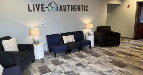 An interior photo of an Ellie Mental Health Therapy Clinic lobby that has stylish gray carpet, blue couches, and the message "LIVE AUTHENTIC" on one wall with the Ellie Mental Health logo.