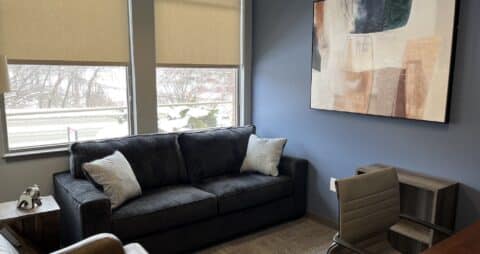 An interior photo of an Ellie Mental Health Therapy Clinic office with two windows half-covered by blinds, a dark blue sofa, a small desk and white desk chair, and a large abstract painting hanging on one wall.