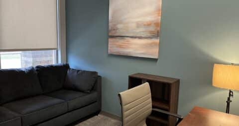 An interior photo of an Ellie Mental Health Therapy Clinic office with dark blue couch, wooden desk with white desk chair, a small floor lamp, a small wooden bookcase, and an abstract painting hanging on one wall.