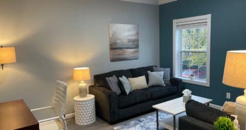 An interior photo of an Ellie Mental Health Therapy Clinic office with a small window, dark blue couch facing a dark blue chair, three floor lamps, a small white table, a desk with a white office chair, and an abstract painting hanging on one wall.