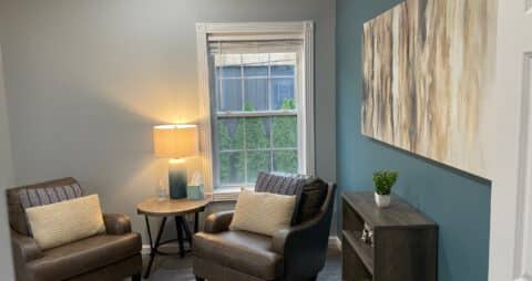 An interior photo of an Ellie Mental Health Therapy Clinic office with small window, two brown chairs, a small table and table lamp, small wooden bookcase, and large abstract painting hanging on the wall.