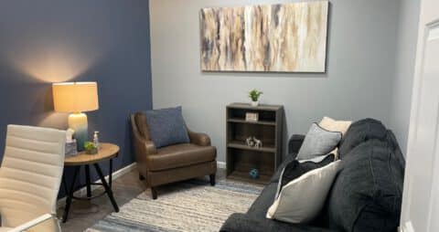 An interior photo of an Ellie Mental Health Therapy Clinic office with a dark blue couch facing a brown leather chair, a small wooden bookcase, a small table with table lamp, a white office chair, and an abstract painting hanging on one wall.