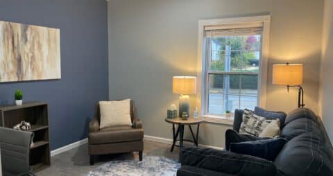 An interior photo of an Ellie Mental Health Therapy Clinic office that has a small window, dark blue sofa, two small lamps, a small wooden bookcase, and an abstract painting hanging on one wall.