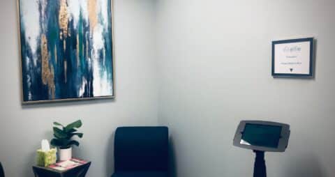 An interior photo of an Ellie Mental Health Therapy Clinic lobby with check in kiosk, blue chair, small plant and Kleenex sitting on top of a small table, an abstract painting hanging on one wall and a framed certificate hanging on the other wall.