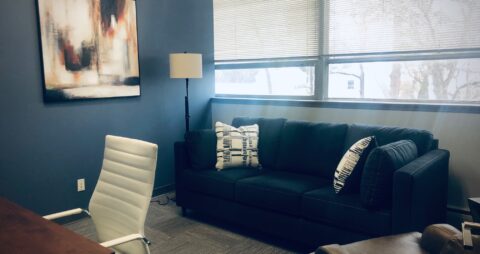 An interior photo of an Ellie Mental Health Therapy Clinic office with a blue couch, lamp, white desk chair and an abstract painting on the wall.