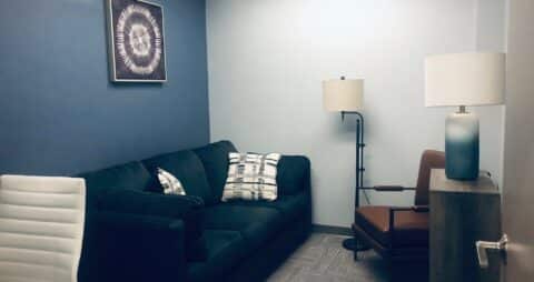 An interior photo of an Ellie Mental Health Therapy Clinic office with a dark blue couch, a brown leather chair, a white office chair, floor lamp, a desk lamp sitting on a small bookcase, and an abstract painting hanging on the wall.