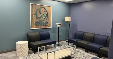 An interior photo of an Ellie Mental Health Therapy Clinic lobby with dark blue and gray chairs, a check-in kiosk, a small white table and a larger glass table, a floor lamp, and the Ellie Mental Health logo hanging on one wall.