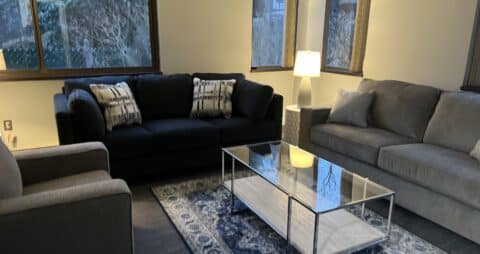 An interior photo of an Ellie Mental Health Therapy Clinic office with gray and dark blue couches, a gray chair, a small glass table and two table lamps.