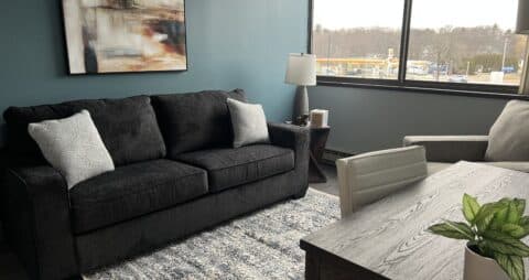 An interior photo of an Ellie Mental Health Therapy Clinic office with dark gray couch, small end table with table lamp, a white office chair sitting in front of a desk, large windows and an abstract painting on the wall.
