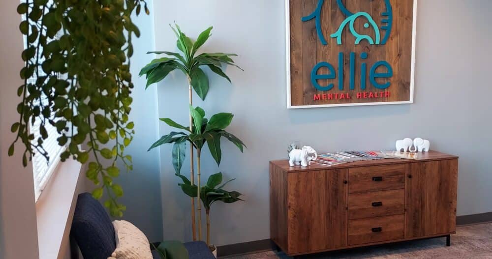 An interior photo of an Ellie Mental Health Therapy Clinic lobby with several plants, two blue chairs, a small wooden desk with white elephant figurines, and the Ellie Mental Health logo hanging on the wall.