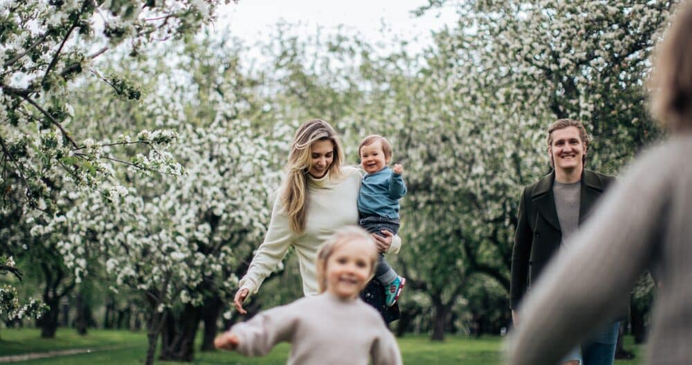 A smiling woman holding her infant son in a park while she and her husband watch their their two kids playing.