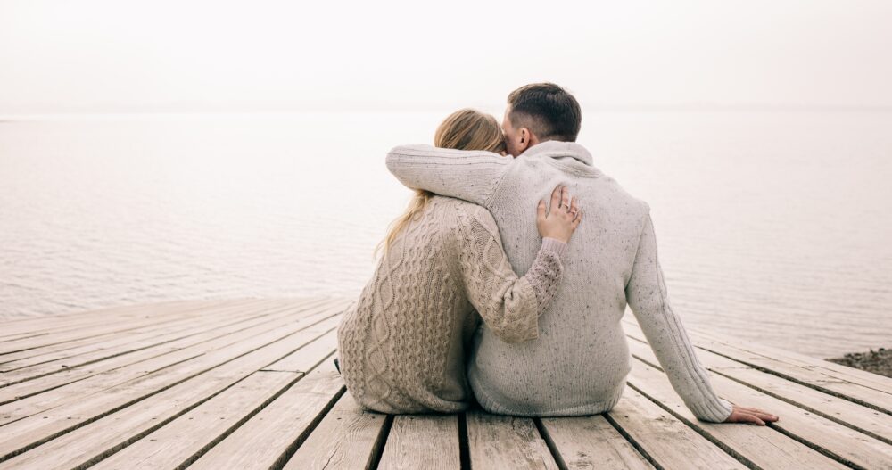 A couple dressed in light beige sweaters embracing while sitting on a dock overlooking the water
