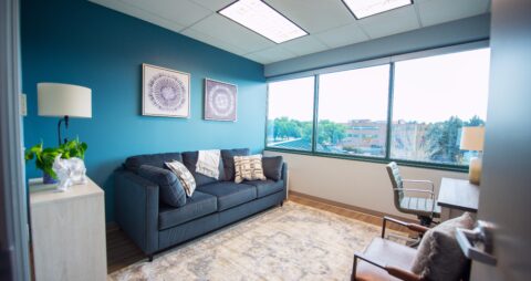 An interior photo of an Ellie Mental Health Therapy Clinic office with three large windows, a dark blue couch, light gray chair, wooden desk with gray desk chair, a table lamp and desk lamp, and two abstract paintings hanging on one wall a