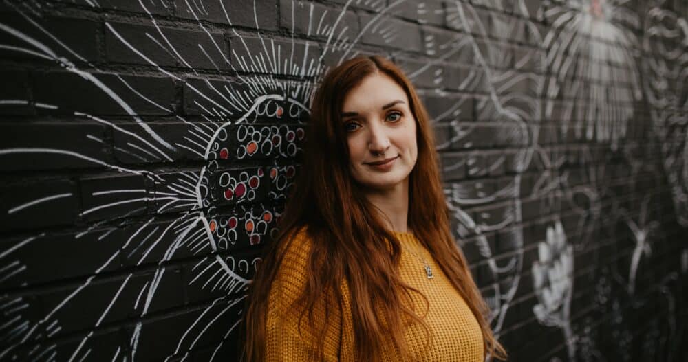 Headshot of a woman with long, copper-colored hair dressed in a yellow sweater standing against a black wall painted with graffiti designs.