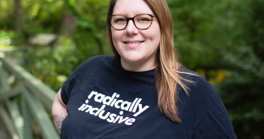 Headshot of a woman wearing dark glasses and tshirt with the logo "radically inclusive"