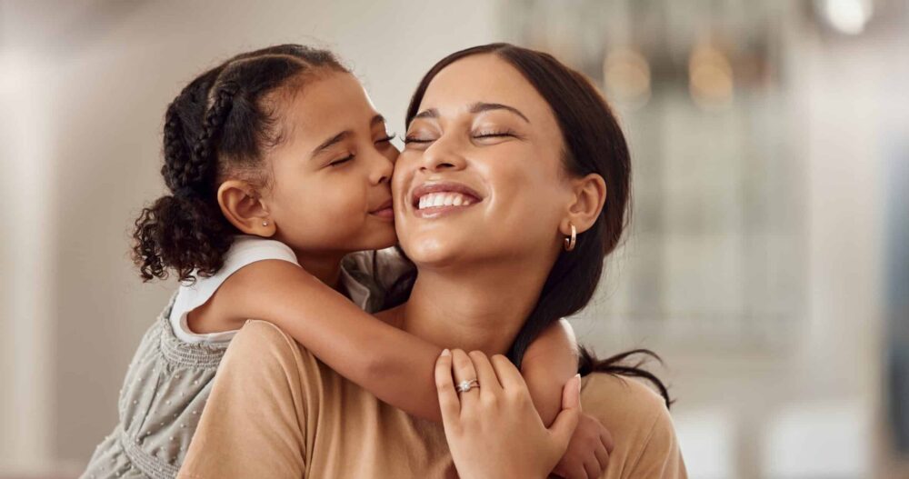 Child hugging her mom from behind and the mom turning her head and smiling at her