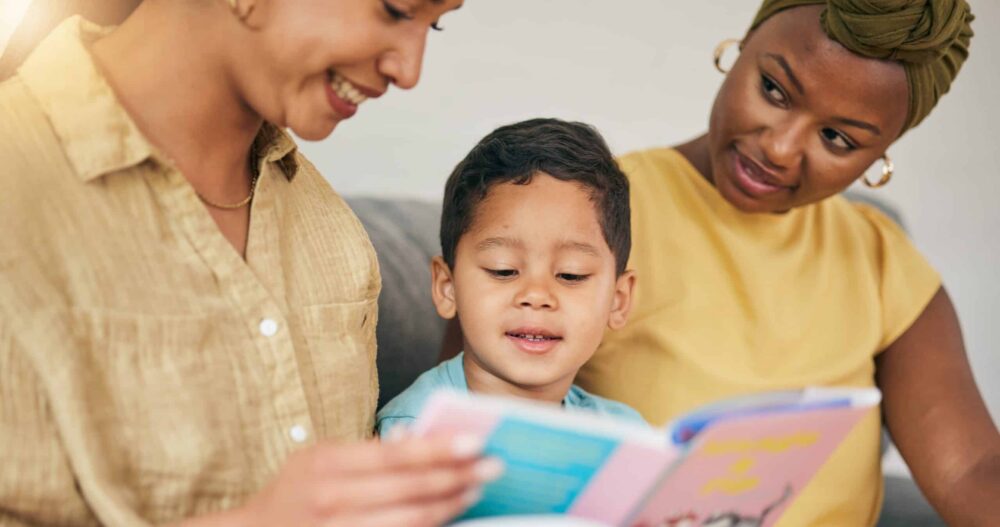 Two moms sitting on the couch with their young child reading a children's book