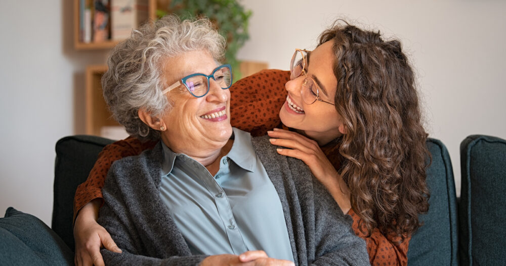 Old grandmother and adult granddaughter hugging at home and looking at each other. Happy senior mother and young daughter embracing with love on sofa. Happy young woman hugging from behind grandma with love.