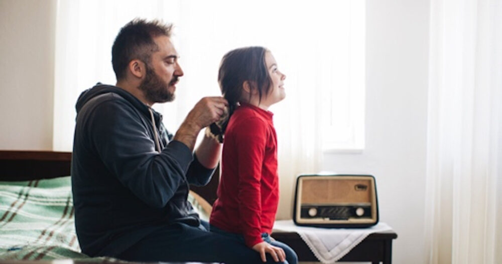 Man sitting on a bed with his young daughter standing in front of him so he can help her with her hair