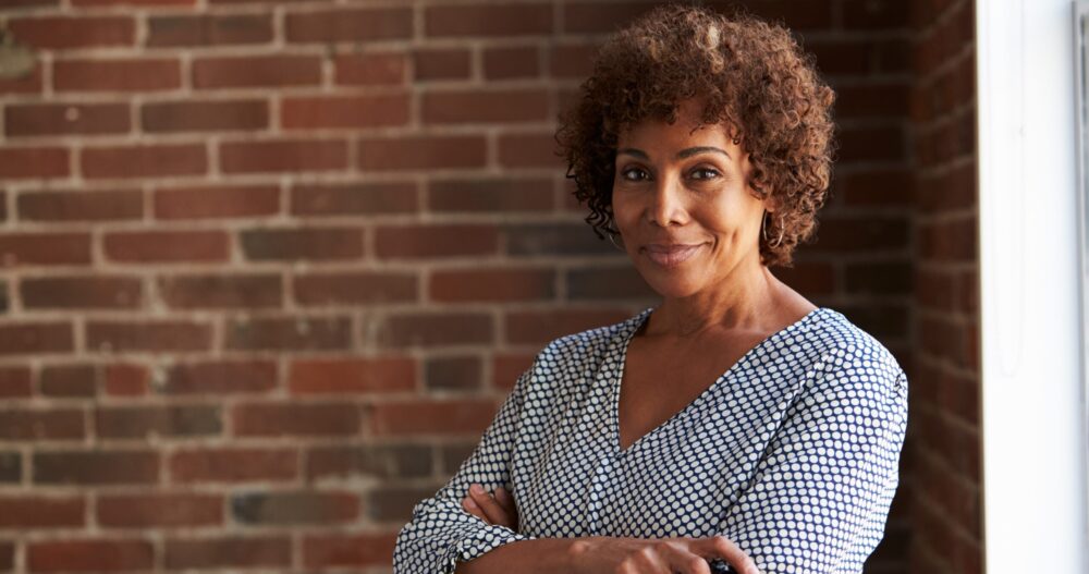 A black woman wearing a white and blue striped shirt standing in front of a brick wall with her arms crossed