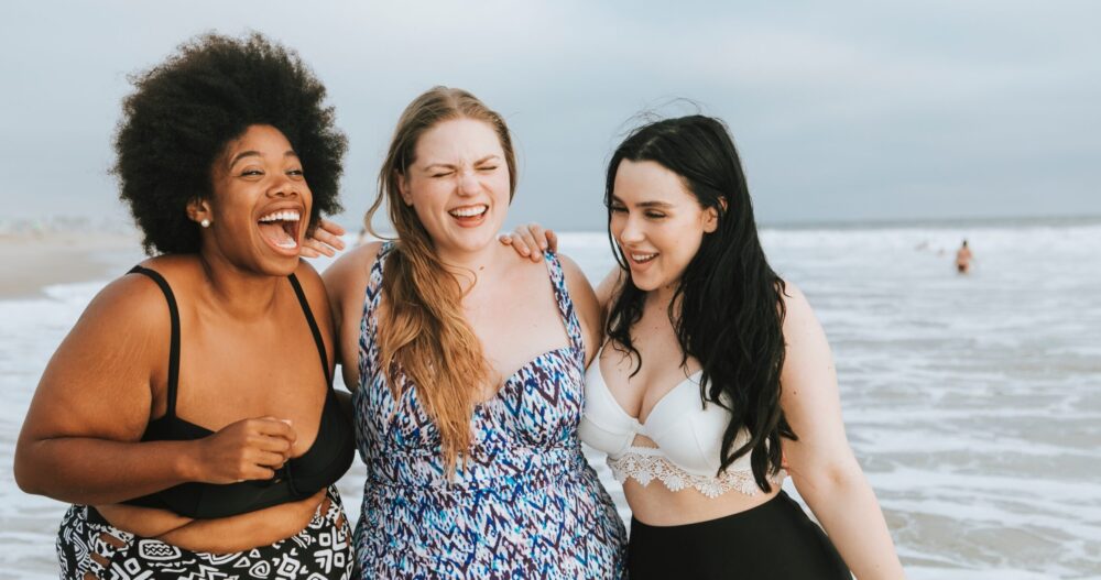 Three women on the beach with the ocean behind them. They are hugging and smiling inf ront of the water
