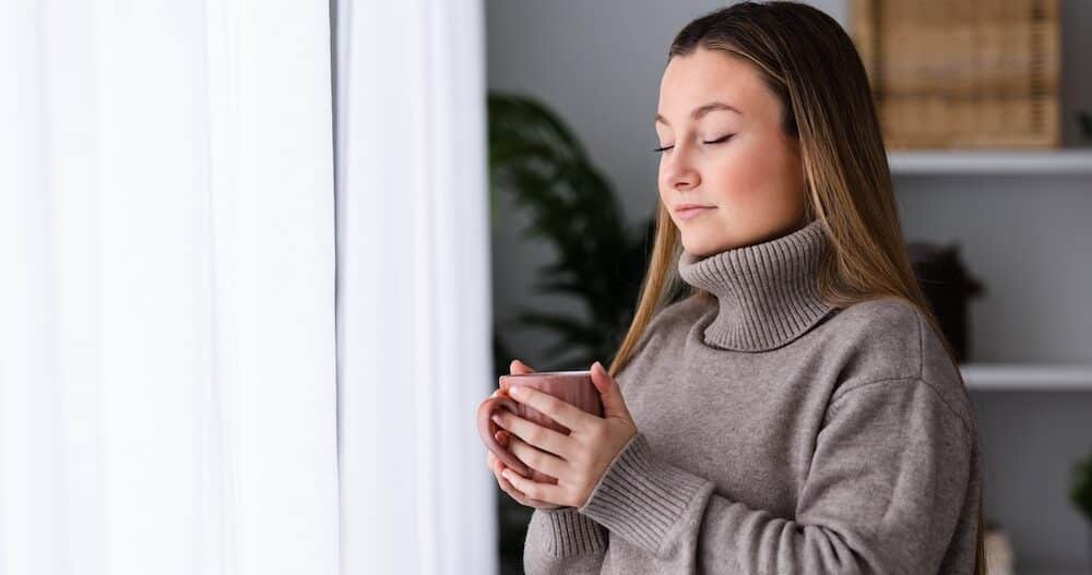 Winter clothing young woman breathing deep beside window while holding cup of hot drink as tea or coffee.