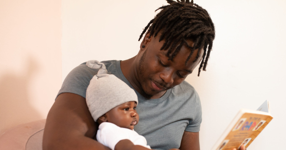 Man holding his young child and smiling at them while holding a book that he is reading to them