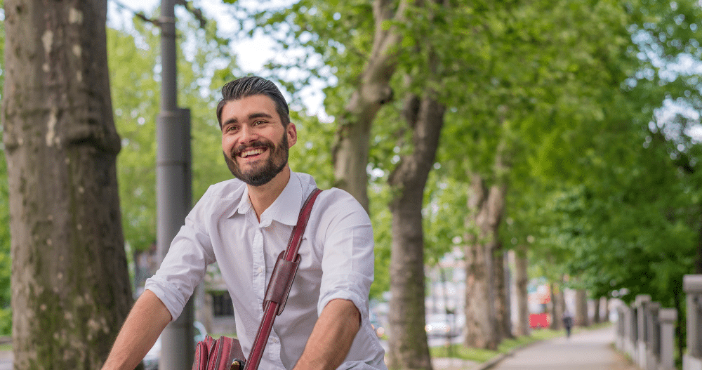 Man with a beard wearing a button up shirt and crossbody bag riding a bike and smiling in front of some trees in the city