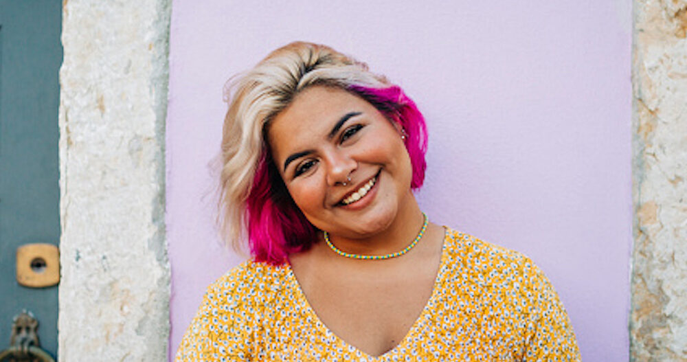 Woman with pink hair standing in front of a colorful wall and smiling