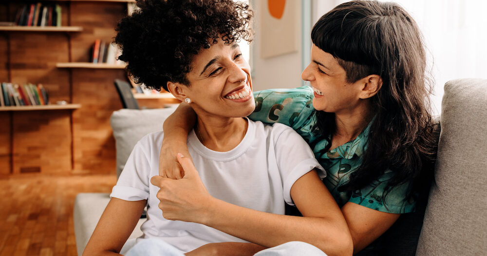 Happy young lesbian couple sharing a laugh while sitting together in their living room.