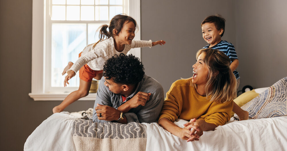 Happy family laughing and having fun together on the bed. Two parents playing with their son and daughter at home. Mom and dad spending quality time with their children on the weekend.