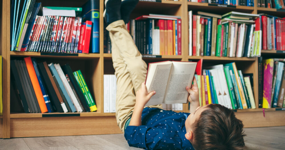 Boy laying on the floor with the feet up, reading a book against multi colored bookshelf in library.
